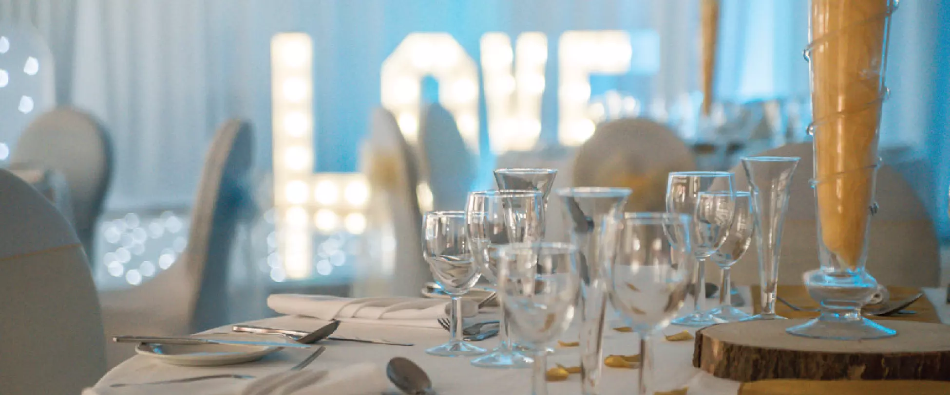 A table set up for a wedding with white table and chair covers, and a yellow centrepiece. In the background, the word LOVE is lit up on the dance floor..